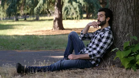 Young pensive man sitting under a tree at park lost in his thoughts Stock Footage 136518632