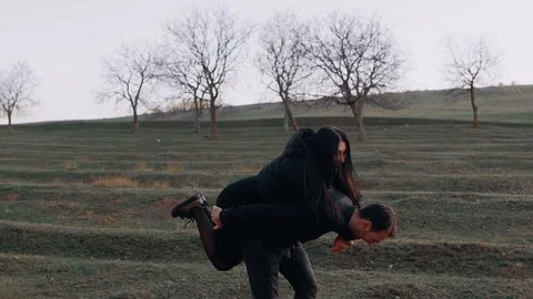 Young people in love fool around on the green meadow at sunset. She jumps behind Stock Footage 154681663