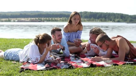 Young people sitting on the green grass and playing cards near the lake. Stock Footage