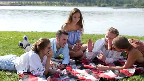 Young people sitting on the green grass and playing cards near the lake. Stock Footage