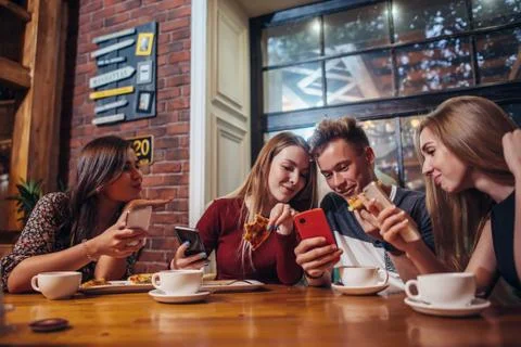 Young people using their mobile phones sitting around the table having a meal in Stock-Fotos