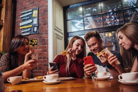 Young people using their mobile phones sitting around the table having a meal in Stock Photos