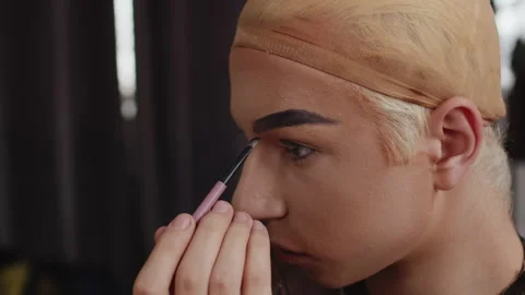 Young performer in drag queen makeup using a brush to fill his eyebrows with Stock Footage 278323565