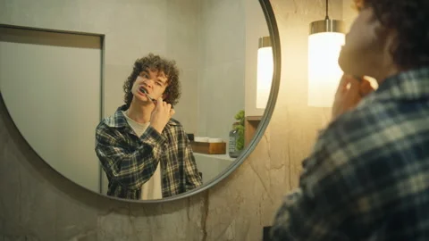 Young person brushes teeth in bathroom while looking at reflection in mirror  Stock Footage 345303464