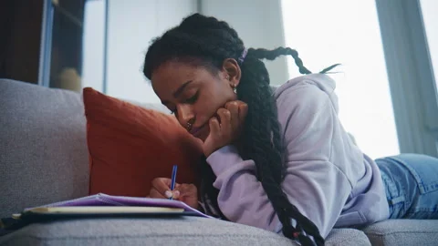 Young person writes in a notebook while lying on a sofa in a cozy indoor space d Stock Footage 339552741