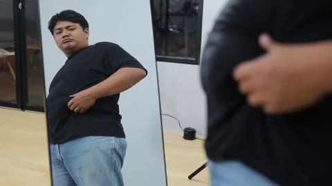 Young plus-size Asian man looking in mirror, feeling unhappy about his body Stock Footage 342283093