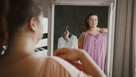Young plus size lady choosing dress at shop, looking at mirror with clothes at Vidéo 143566913