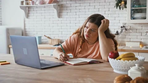 Young plus size lady making notes, watching online webinar at home, kitchen Stock Footage 143568211