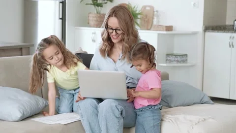 Young positive mother trying to work on laptop with children at home Stock Footage 152664293