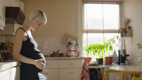 Young pregnant mother standing in her kitchen rubbing her baby bump, in slow mot Stock Footage