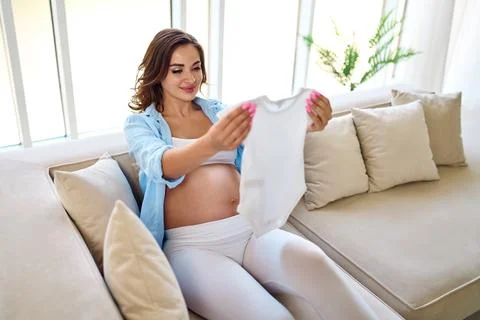 Young pregnant woman sitting on the couch and looking at the little baby clot Stock Photos