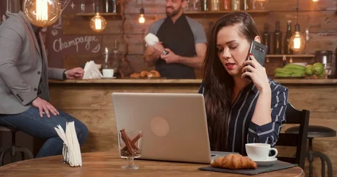 Young pretty lady talking at her phone while working at her laptop remotely Stock Footage 103373217