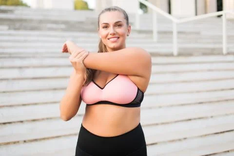 Young pretty smiling plus size woman in pink sporty top and leggings stretching Stock Photos