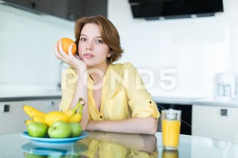 Young pretty woman holding fruits sitting near desk in the kitchen Stock Photos