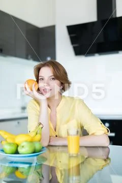 Young pretty woman holding fruits sitting near desk in the kitchen Stock Photos