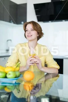 Young pretty woman holding fruits sitting near desk in the kitchen Stock Photos