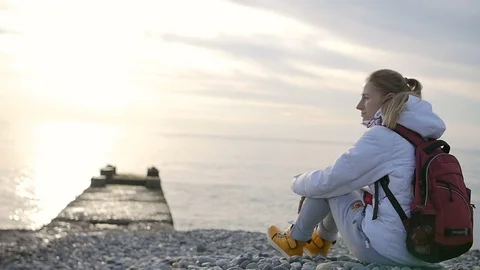 Young pretty woman in jacket with backpack sitting near the sea at sunset Video stock 71629239