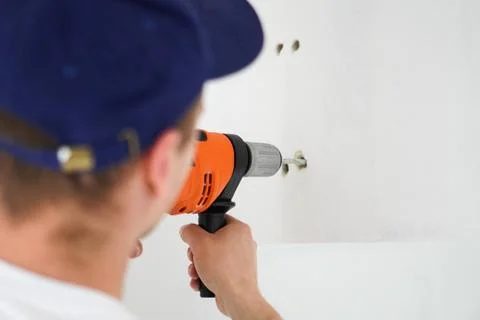 Young professional worker with drill making a hole in white wall. Stock Photos