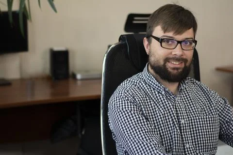 Young programmer man in glasses with a beard sits at a table on a chair in a Stock Photos