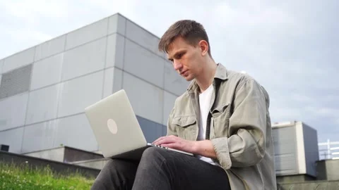 A young programmer works at his laptop outside the office. Cityscape. Stock Footage 245665616