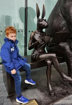Young red-haired boy in blue jacket sits beside whimsical bronze rabbit Stock Photos