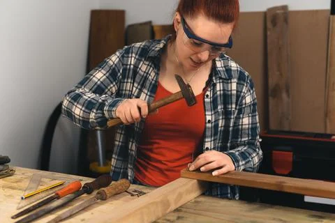 Young red-haired carpenter hammering a nail into a wooden board in her small Stock Photos
