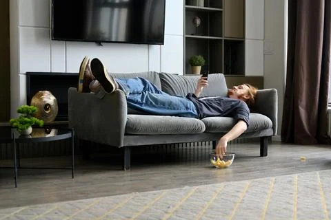 A young red-haired guy is lying on the sofa and eating corn flakes Stock Photos