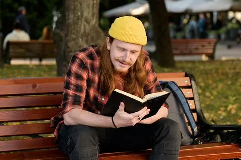 A young red-haired guy sits on a bench and reads a book Stock Photos
