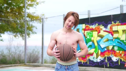 Young red-haired guy without t-shirt on sports ground holds basketball in hand Vidéo 239564599