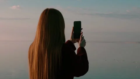 Young red-haired woman capturing a serene lake view on her smartphone, tranquil  動画素材 303395996