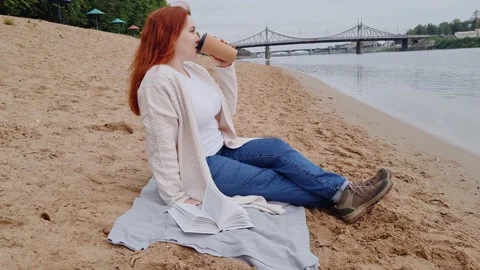 Young red-haired woman drinking coffee on beach. Enjoy autumn cloudy day Stock Footage 187546031