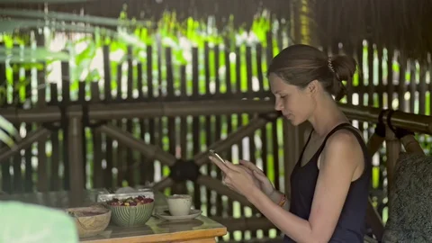 Young red haired woman using smartphone and drinking tea at table in island cafe Stock Footage 124448989
