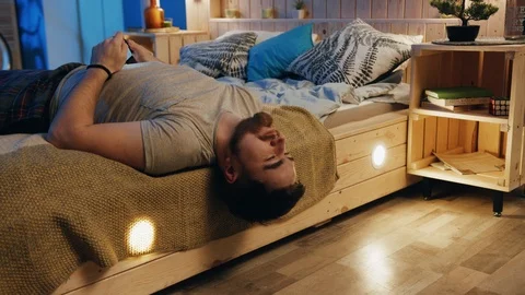 Young relaxed man hanging from bed, holding smartphone in hand, surfing social Stock Footage 105653656