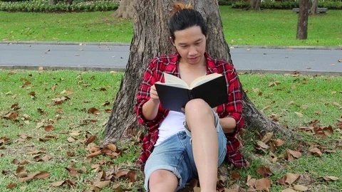 Young relaxed man is reading a book under tree at the park outdoors Stock Footage 79985221
