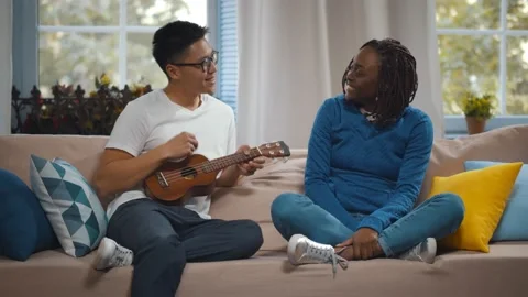 Young romantic multiethnic couple playing ukulele on couch in living room Stock Footage 140348612