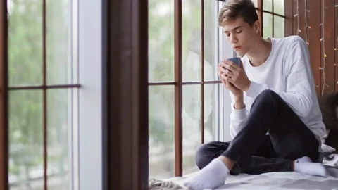 A young sad guy sits by the window and drinks a hot drink. Stock Footage 154715847
