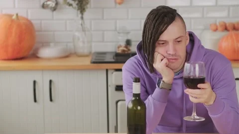 Young sad man with dreadlocks looks at glass of red wine, sitting at table in Stock Footage 220509236