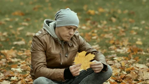 A young sad man in a hat sits among the fallen leaves, looking at a yellowed Stock Footage 160651230