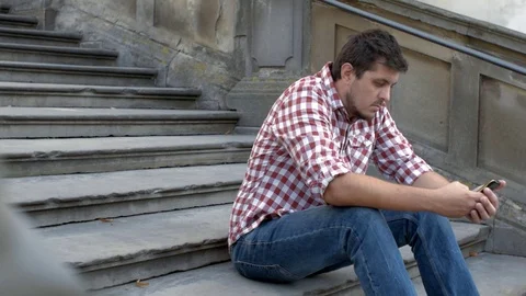 A young sad man is sitting on the old stairs and using app on the smartphone Stock Footage 79495429