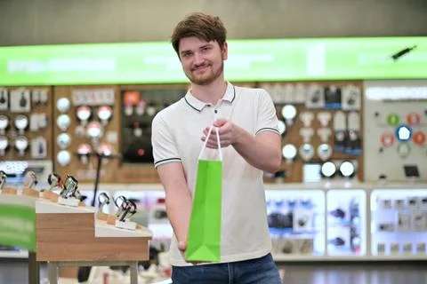 Young salesman in electronics store puts goods into paper bag and holds out Stock Photos