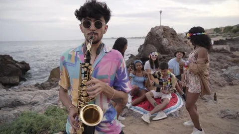 Young saxophonist looking at camera plays at group of friends party on beach Stock Footage 275560537