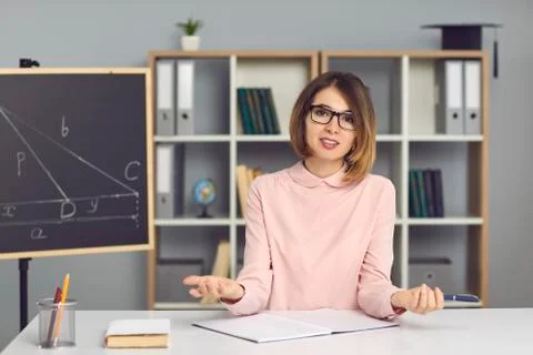 Young school math teacher sitting at desk, explaining new topic and looking at Stock Photos