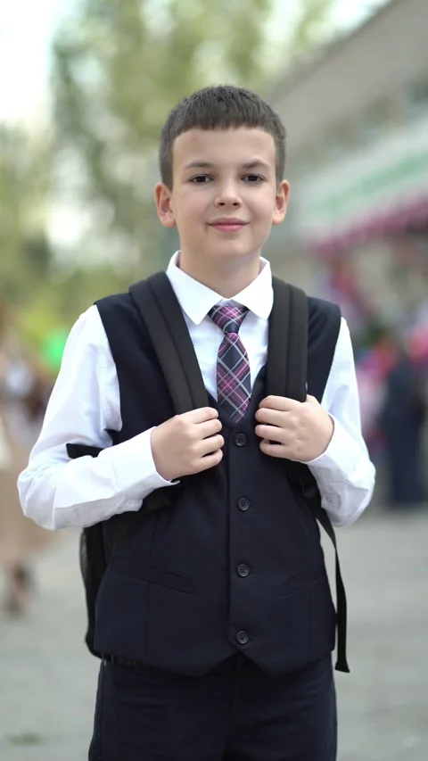 Young schoolboy holds backpack straps, posing outdoors by school Stock Footage 344312751