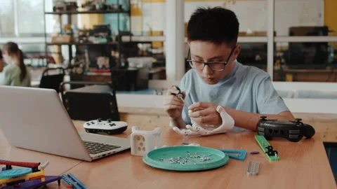 Young schoolboy sitting at the table with a laptop and assembling small parts of Stock Footage 242170566