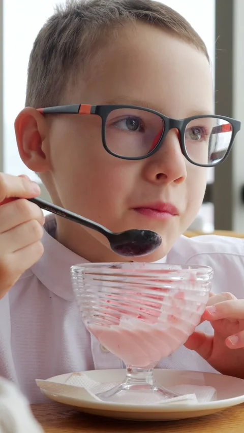 Young schoolboy wearing eyeglasses eats pink dessert using spoon indoors. Happy Stock-Footage 346407342