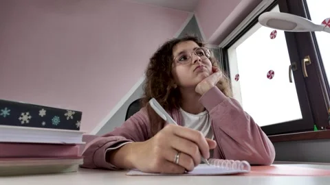 Young schoolgirl thinking while doing homework at desk near window Stock Footage 328420687