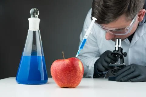 Young scientist using blue liquid, apple, syringe and microscope. Stock Photos