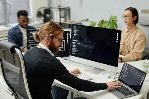 Young serious diversity programmer typing on laptop keyboard by workplace Stock-Fotos