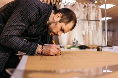 Young serious tailor with beard looking at drawing near wooden table with thr Stock Photos