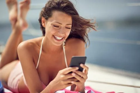 A young sexy girl is texting on a smartphone while is enjoying a sunbath. Sum Stock Photos